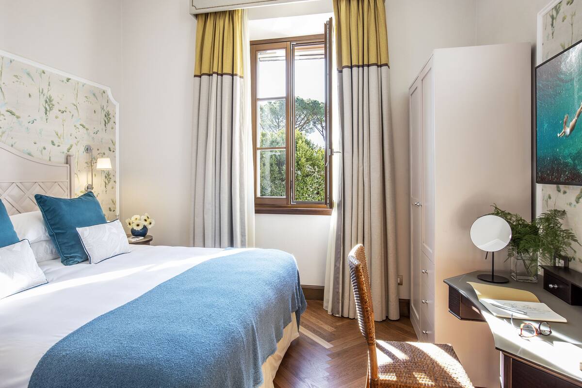 Ein Schlafzimmer mit einem Bett, einem Schreibtisch, einem Stuhl, einem Fenster mit Vorhängen und einem wandmontierten Spiegel.