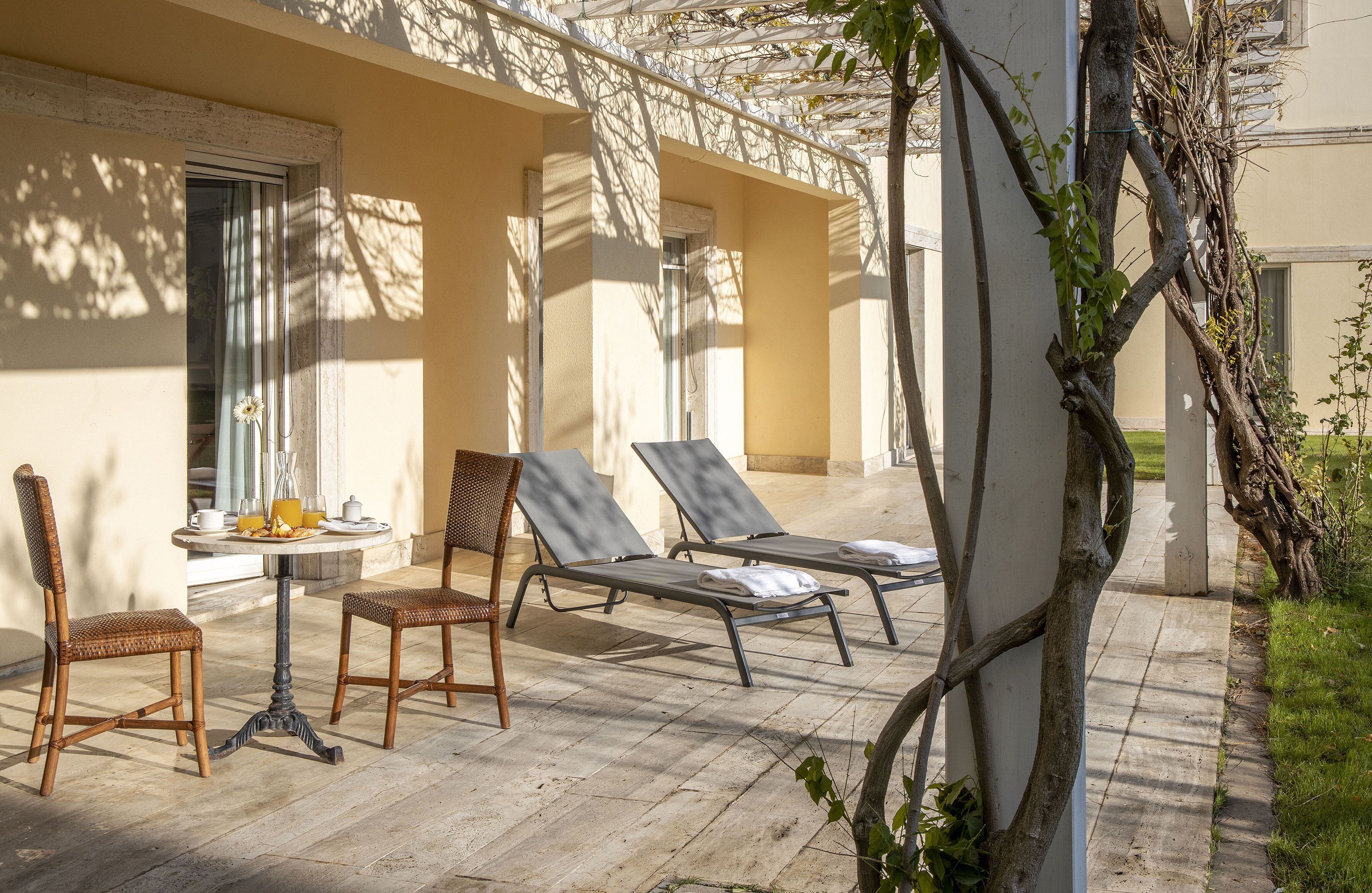 A sunlit outdoor seating area with wicker chairs and lounge chairs, a table with breakfast items, and a pergola with climbing vines.