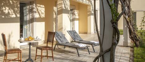 A sunlit outdoor seating area with wicker chairs and lounge chairs, a table with breakfast items, and a pergola with climbing vines.