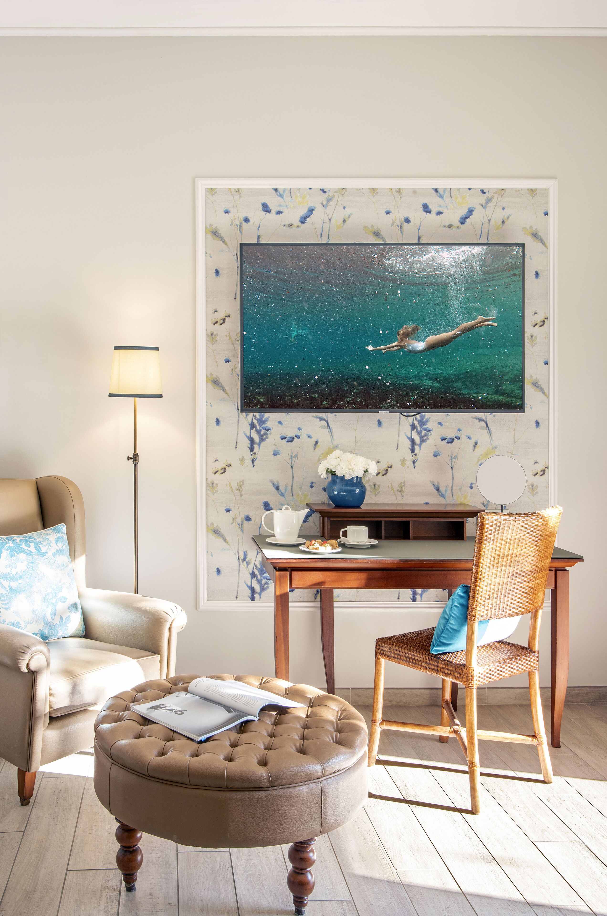 A room with a beige sofa, a round ottoman with a book, a wicker chair, a wooden table with a vase, and a wall-mounted TV displaying a mermaid.