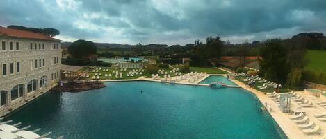 A large swimming pool with lounge chairs and umbrellas, surrounded by buildings and greenery.