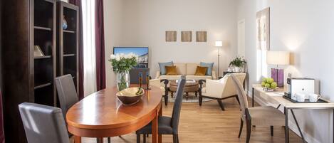 A modern living room with a wooden dining table, grey upholstered chairs, a sofa with blue cushions, a coffee table, and a TV in the background.