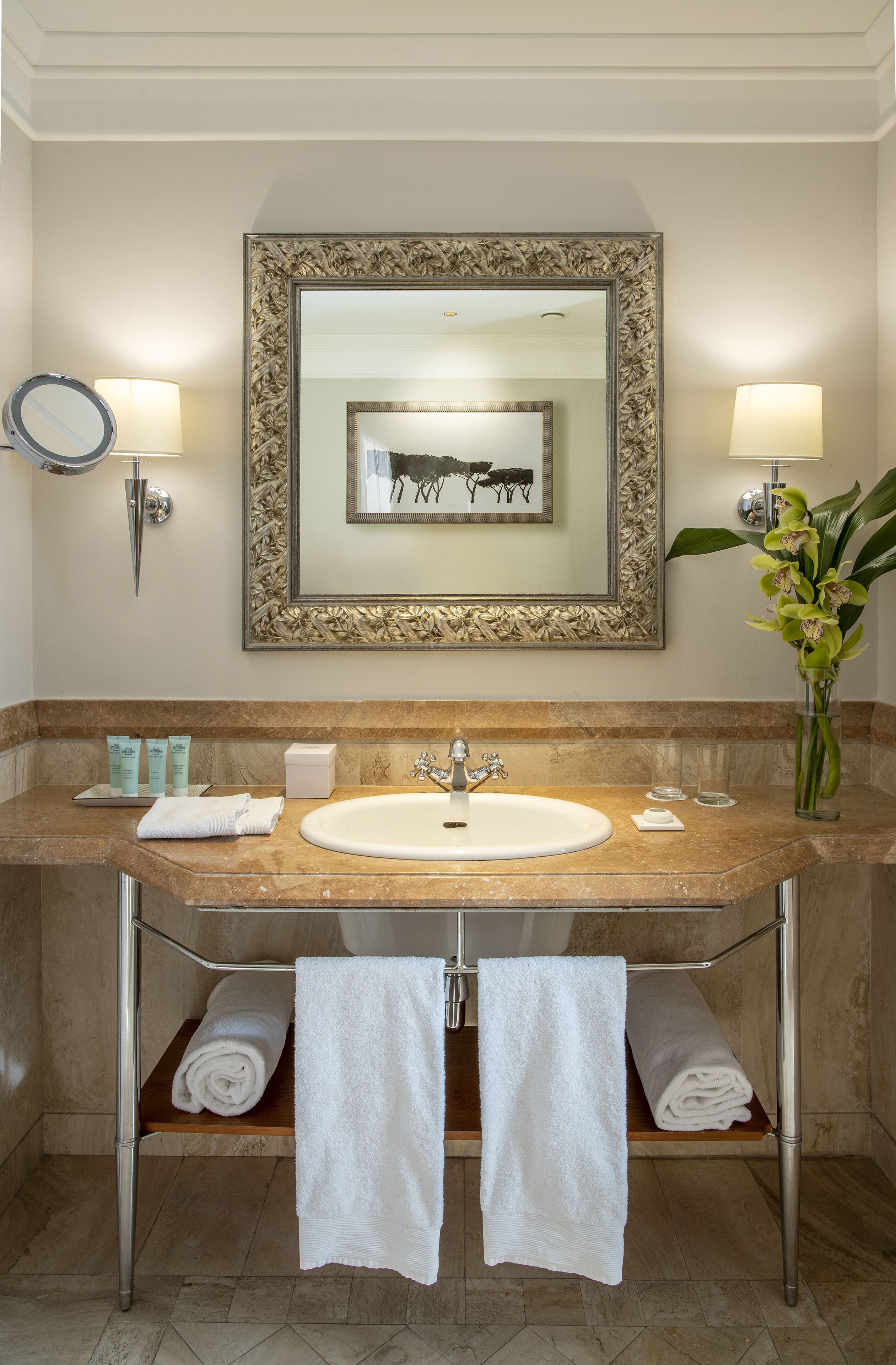 A bathroom with a marble sink, two wall-mounted lamps, a framed artwork, a vase with flowers, and two towels on a shelf.