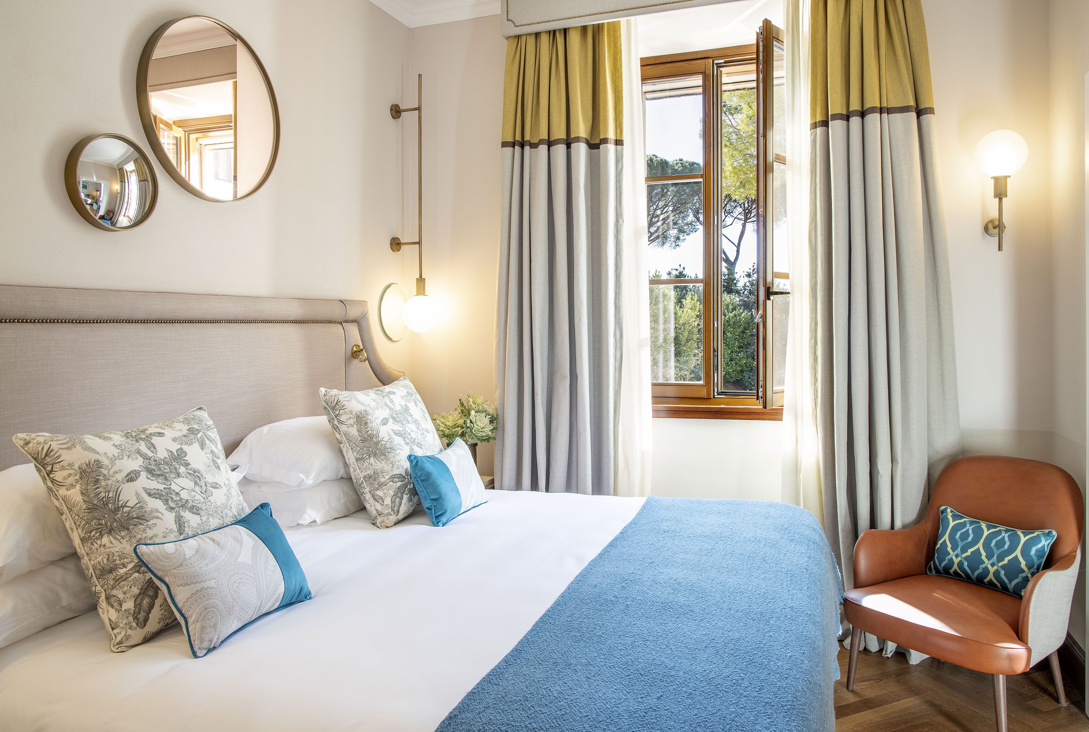 A neatly made bed with a blue blanket, a chair with a patterned cushion, and a window with curtains.