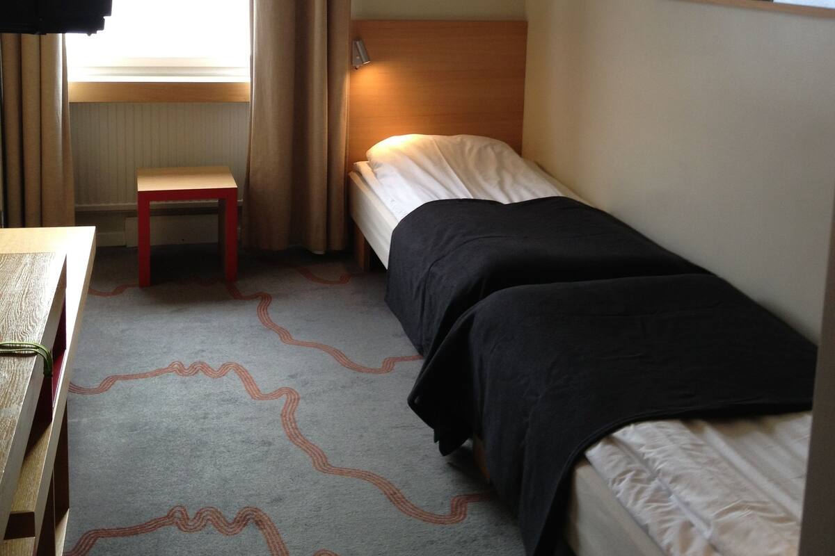 A small hotel room with a single bed, a bedside table, a chair, a small table, a window with curtains, and a wall-mounted picture.