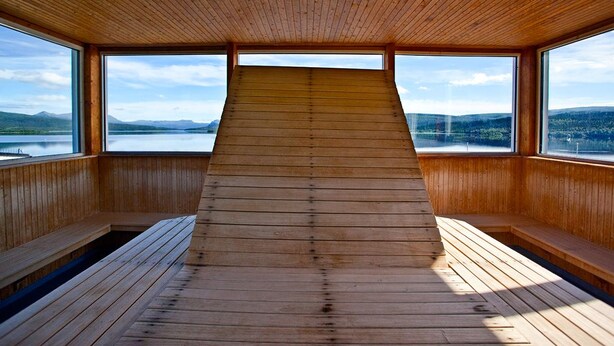 Un interior de madera con techo inclinado y amplias ventanas que ofrecen vista a un lago y montañas.