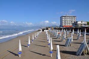 A beach with umbrellas and sun loungers, a building with multiple floors, and a view of the sea and mountains in the background. - Riviera Spineta (Battipaglia)