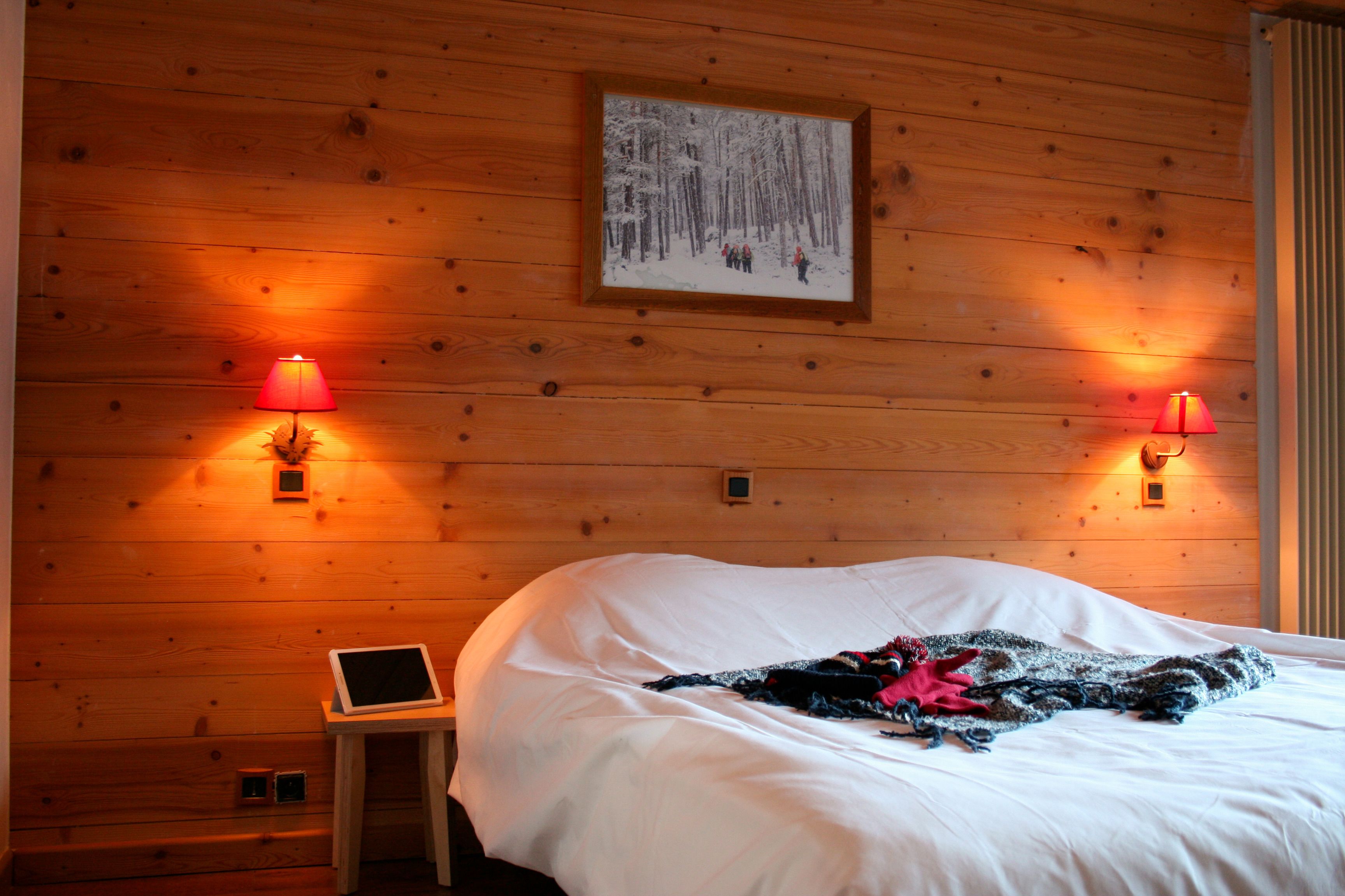 Una habitación con paneles de madera, una cama, dos lámparas fijadas a la pared, un cuadro enmarcado y una pequeña mesa con una tableta.