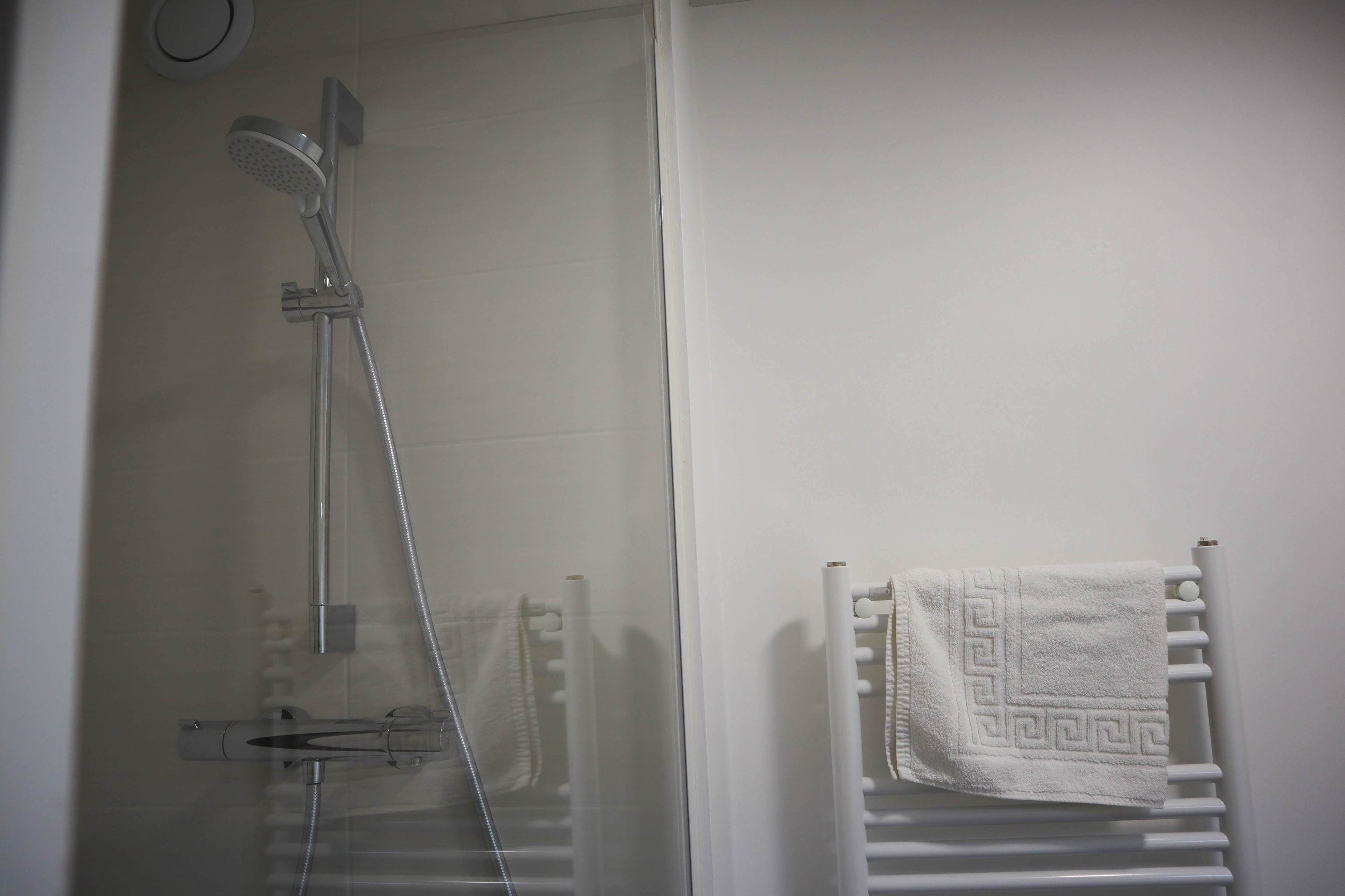 A shower with a handheld showerhead and a wall-mounted faucet, a towel rack with a folded towel, and a white tiled wall.