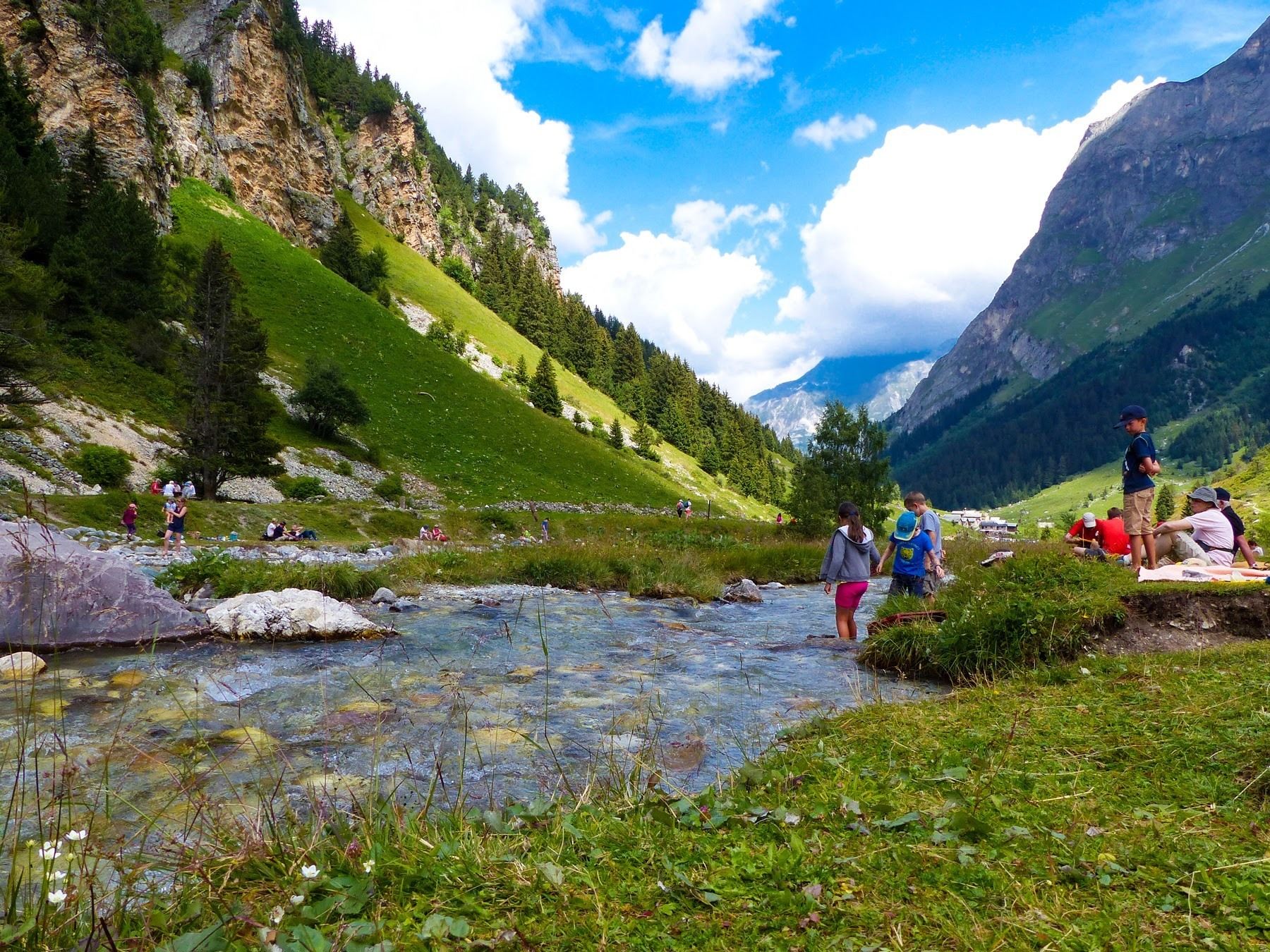 A mountainous landscape with a clear stream, lush greenery, and people enjoying the outdoors.