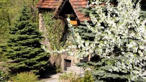 A stone cottage with a tiled roof, surrounded by lush greenery and a stone pathway leading to a bench. - Hotel Przy Oślej Bramie - Zamek Książ (Walbrzych)