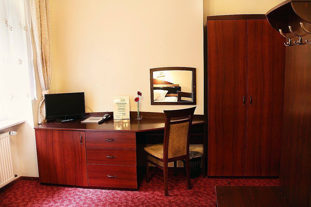 A hotel room with a wooden desk, a chair, a mirror, a wardrobe, and a radiator.