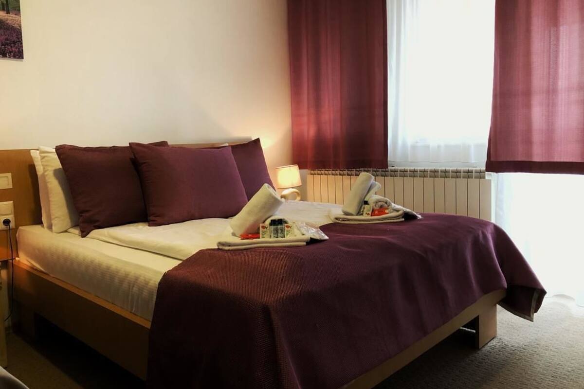 A neatly made bed with a white and purple bedspread, a bedside table with a lamp, and a window with curtains.