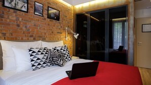 Une chambre avec un mur en briques, un lit recouvert de draps blancs, des oreillers à motifs noir et blanc, une couverture rouge, un ordinateur portable posé sur le lit et une porte-fenêtre coulissante donnant sur une pièce sombre.
