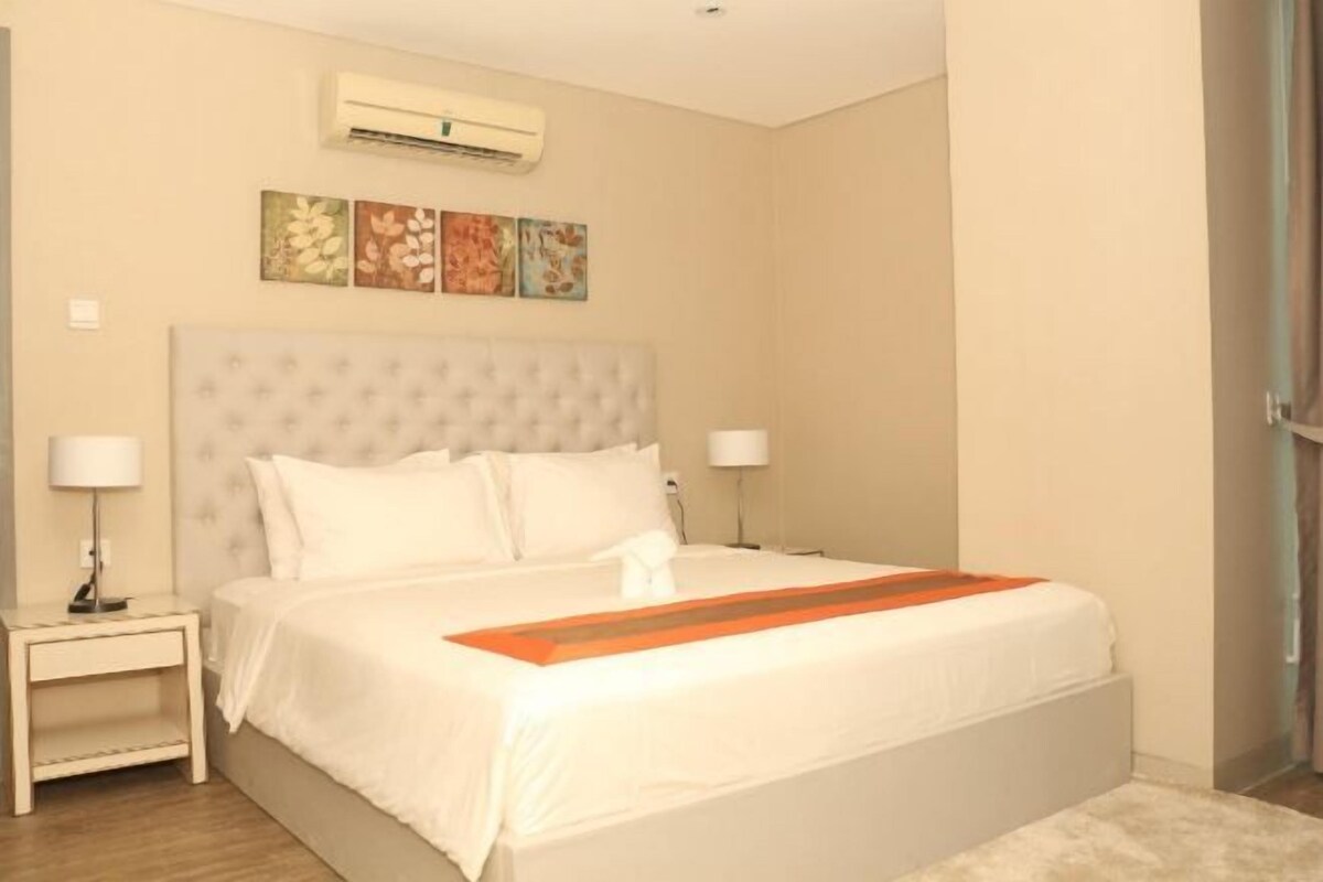 A hotel room with a large bed, two bedside tables, an air conditioning unit, and three wall-mounted artworks.