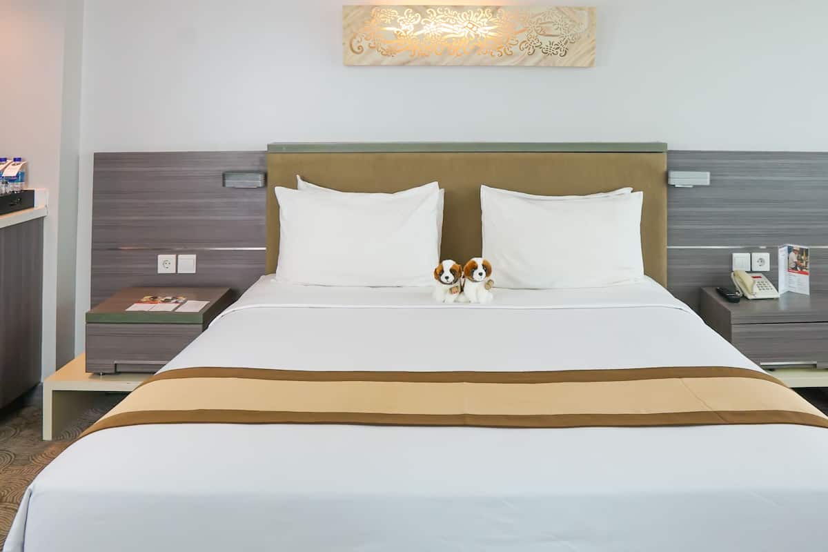 Hotelkamer met een groot bed, twee witte kussens, een bruin hoofdeinde en een nachtkastje met een telefoon en een brochure.