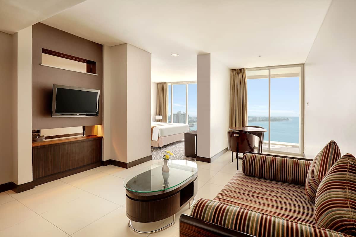 Een moderne hotelkamer met een groot bed, een televisie, een glazen salontafel, een gestreepte bank en uitzicht op de stad en het water.