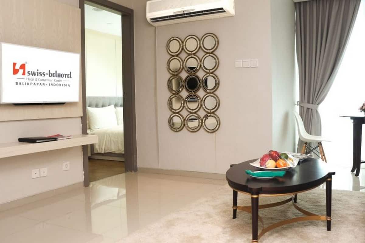Een moderne hotelkamer met een flatscreen-tv waarop het Swiss-Belhotel logo staat, een wandspiegelopstelling, een ronde tafel met een schaal fruit, en een groot raam met gordijnen.