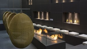 A modern interior with hanging wicker chairs, a glass-top table with a built-in fire feature, and shelves with bottles.
