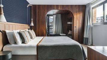 A hotel room with a wooden headboard, a large bed with a grey blanket, a small round table, and a view of the outside through a window.