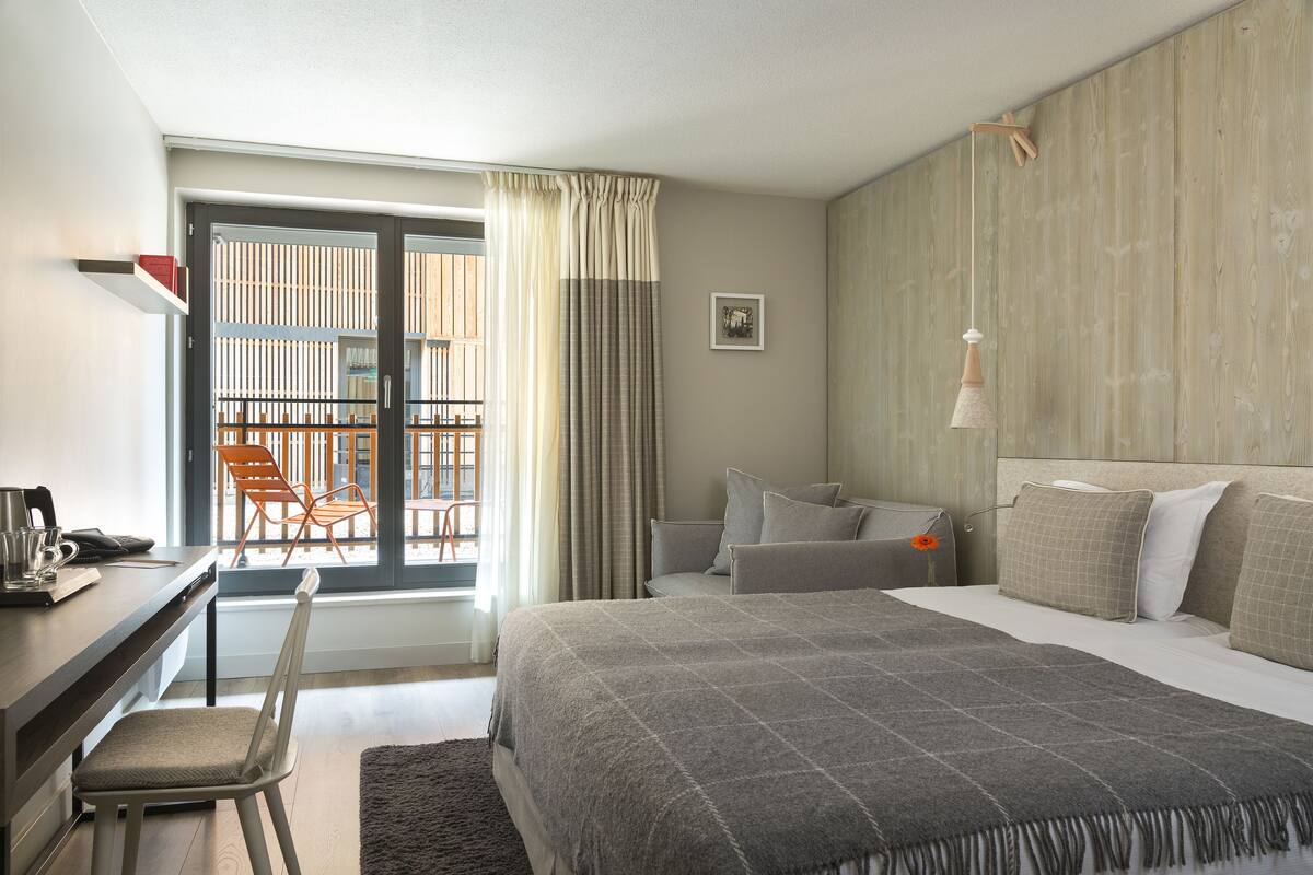 A hotel room with a bed, a desk, a chair, a sofa, and a balcony with chairs.