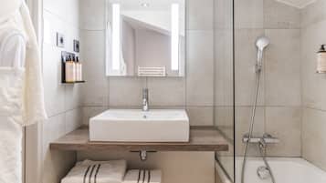 Une salle de bain moderne avec un lavabo blanc, un miroir et une douche. On y trouve des serviettes et des articles de toilette sur le comptoir.