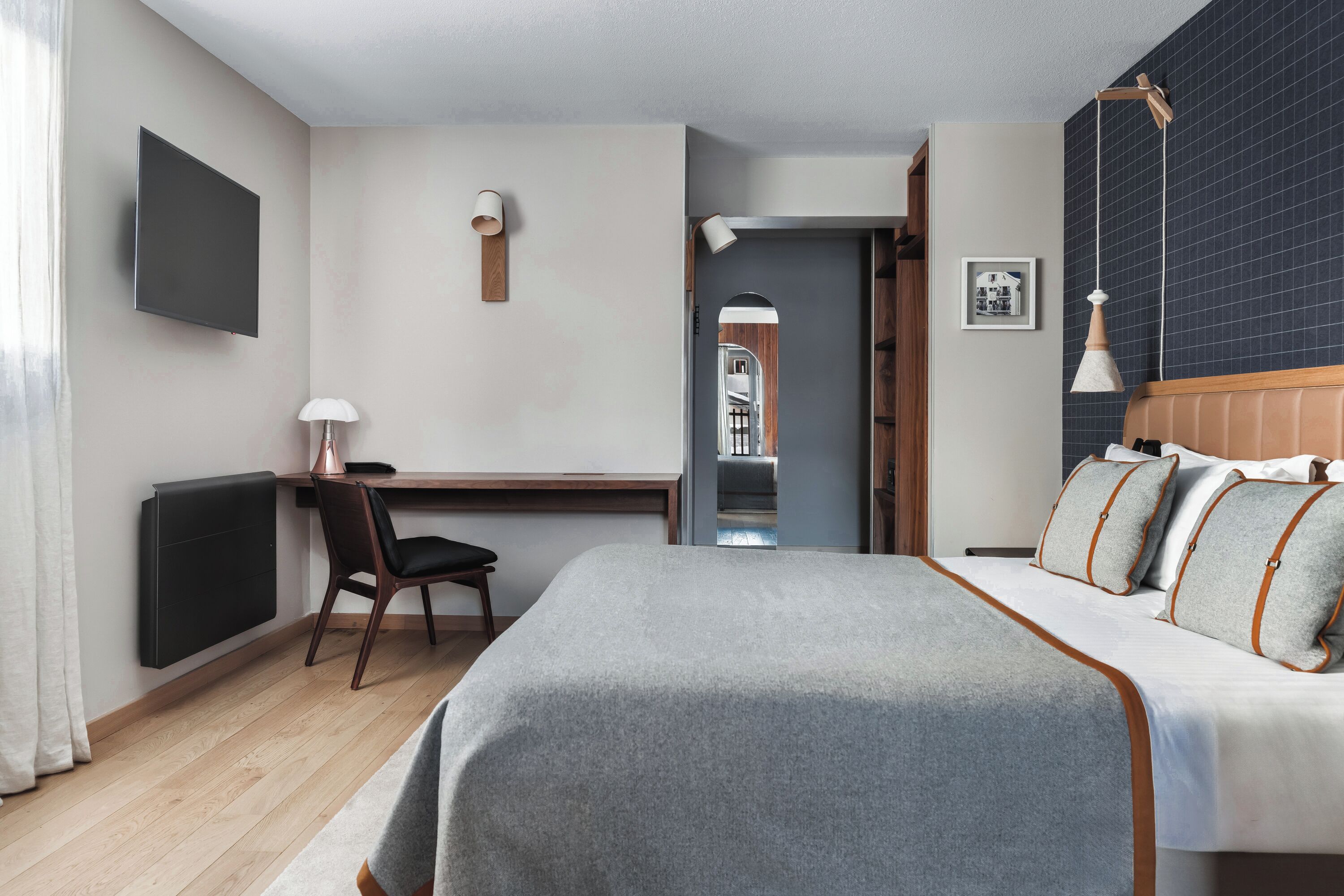 A modern hotel room with a bed, desk, chair, and a view of the hallway.