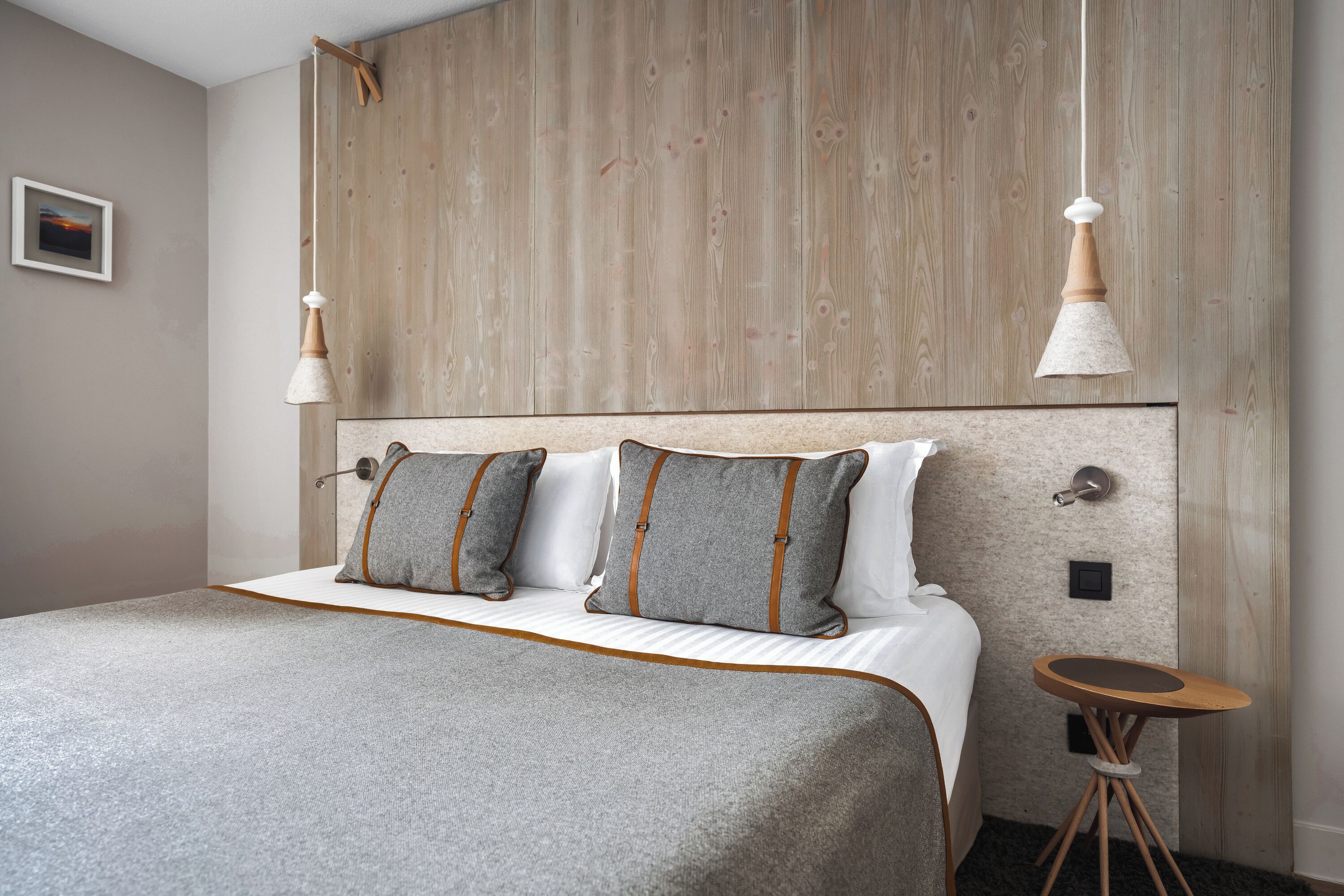 Una cama bien tendida con edredón gris y blanco, dos almohadas con detalles marrones y una mesita de noche de madera.
