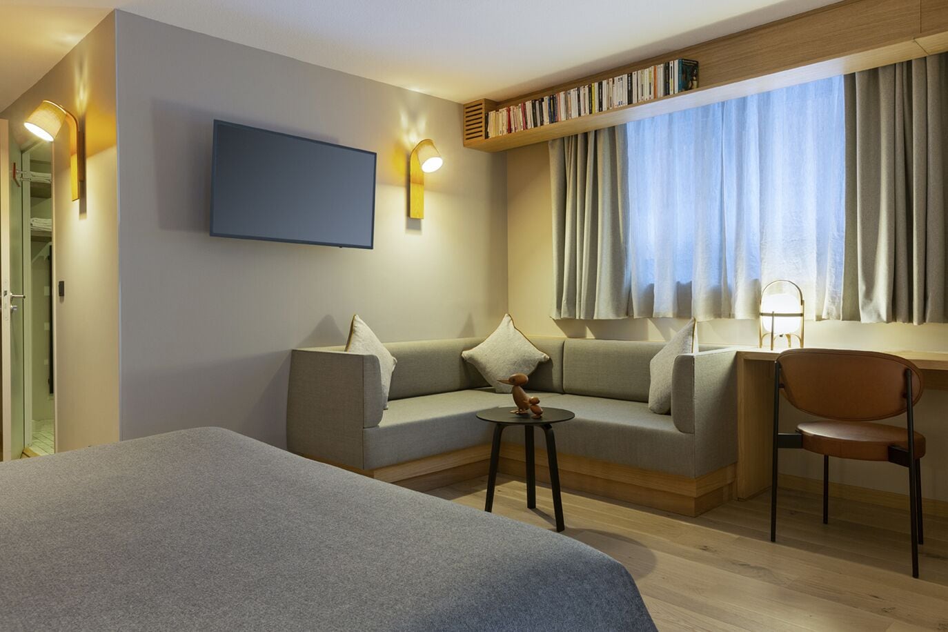 Une chambre d’hôtel moderne équipée d’un lit, d’un canapé, d’un bureau et d’une chaise. Un téléviseur est fixé au mur et une bibliothèque contient des livres.