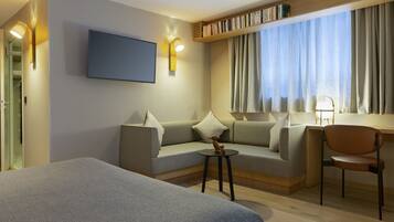 Une chambre d’hôtel moderne équipée d’un lit, d’un canapé, d’un bureau et d’une chaise. Un téléviseur est fixé au mur et une bibliothèque contient des livres.