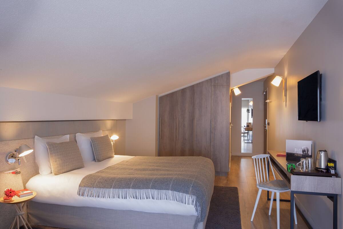 A modern hotel room with a large bed, bedside table, lamp, and a desk with a chair.