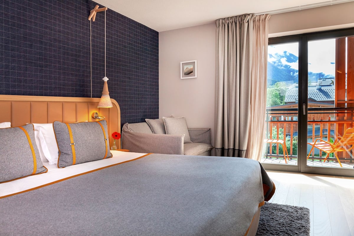 A modern hotel room with a large bed, a seating area with a sofa and armchairs, a view of a balcony with outdoor furniture, and a large window with curtains.