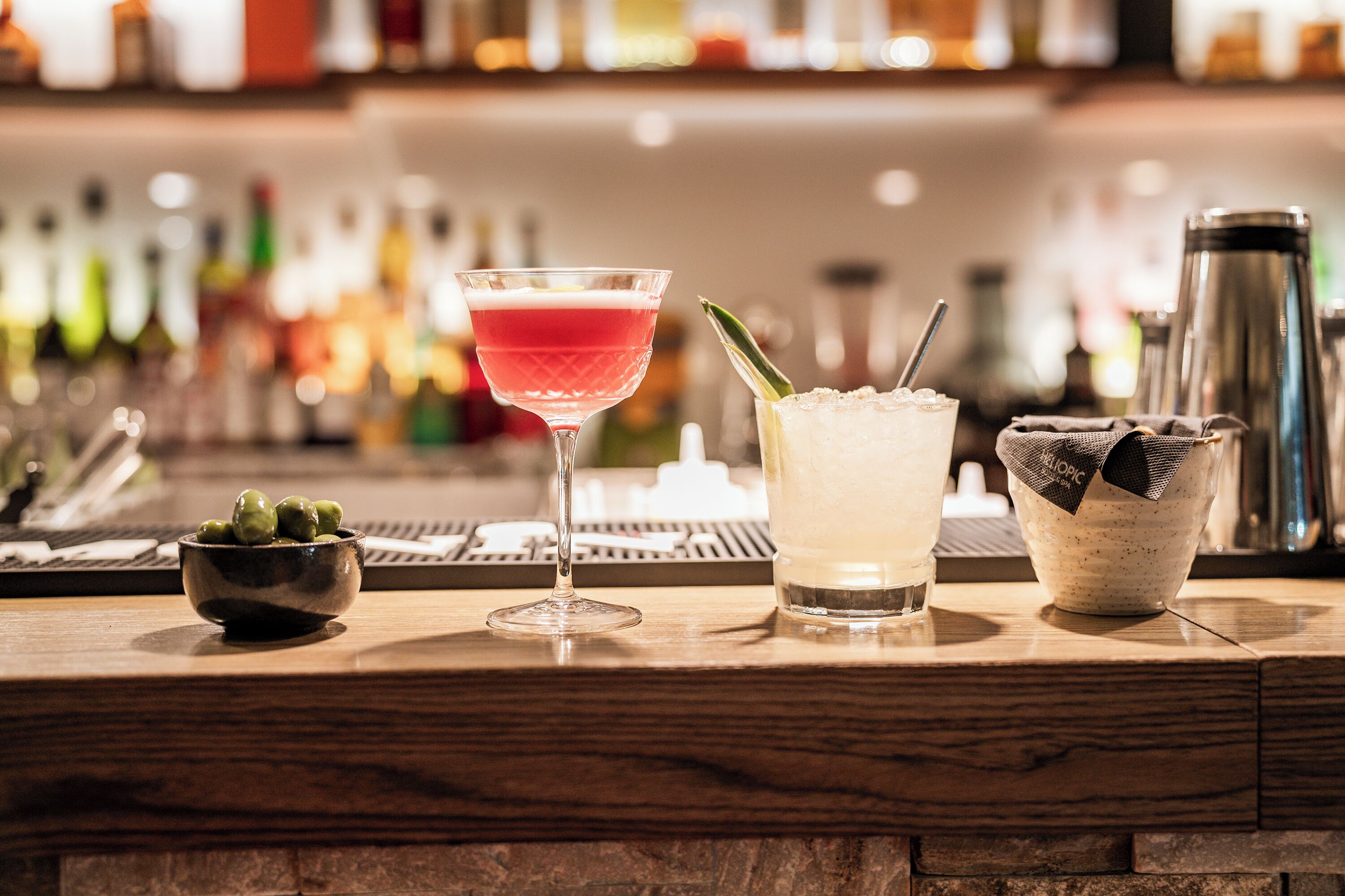 Una barra con una copa de martini, un cóctel con pajita y un bol de aceitunas.