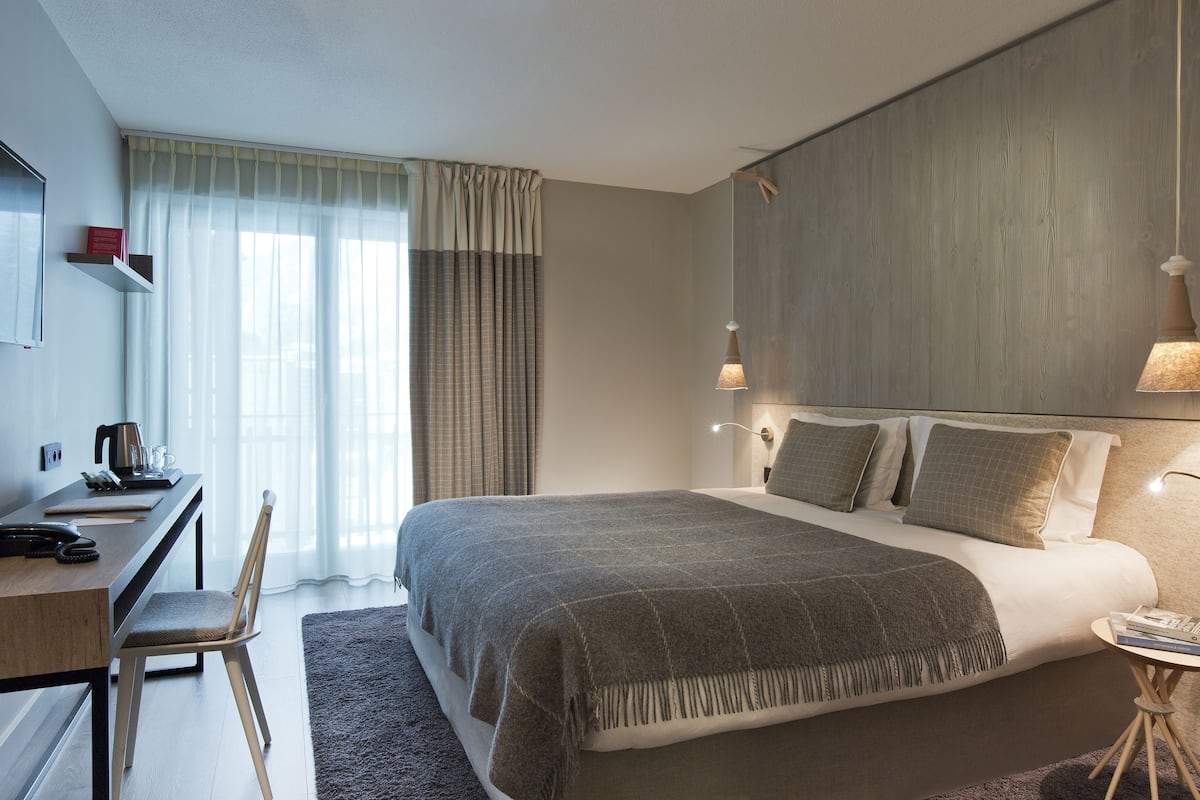 A modern hotel room with a large bed, a desk with a chair, a small table, and a nightstand.