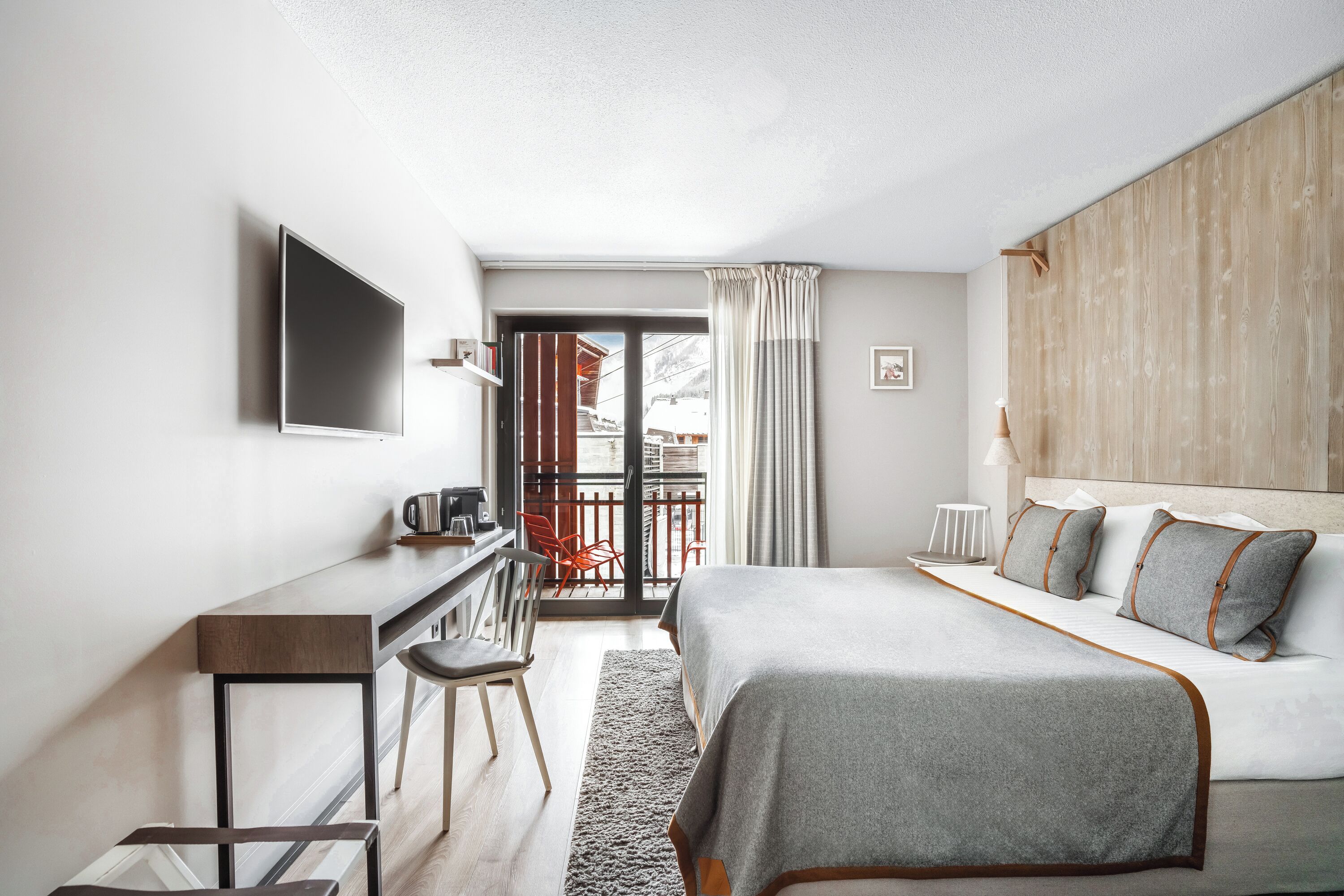 A modern hotel room with a bed, desk, and chair, a TV mounted on the wall, and a view of a balcony with chairs.