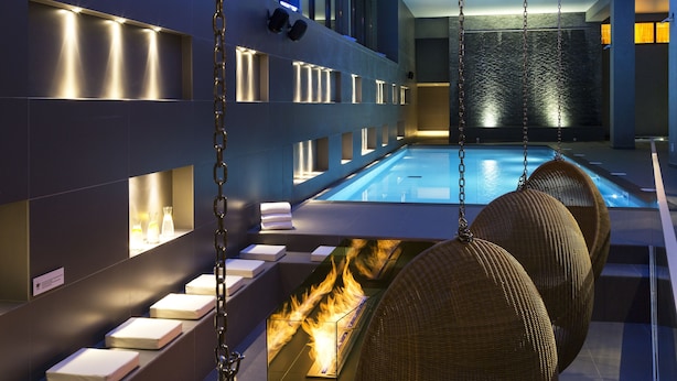 A modern indoor pool area with a glass barrier, wicker chairs, and a fire feature.