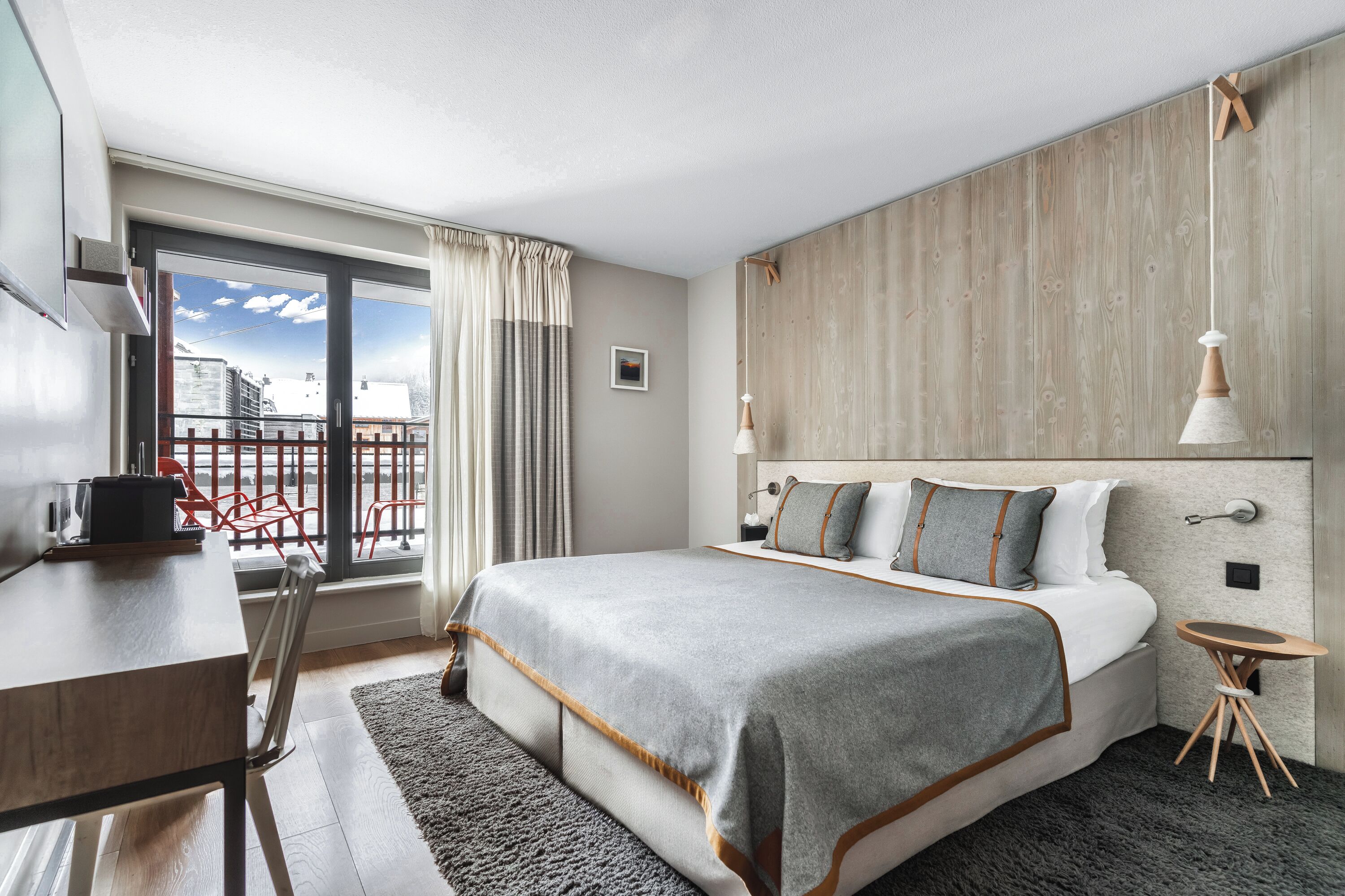 Une chambre d’hôtel moderne avec un grand lit, un bureau et un balcon agrémenté de chaises rouges.