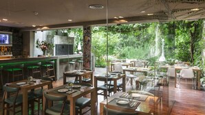 A restaurant with wooden tables and chairs, a bar area, and large windows offering a view of greenery. - Home Hotel Buenos Aires (Buenos Aires)