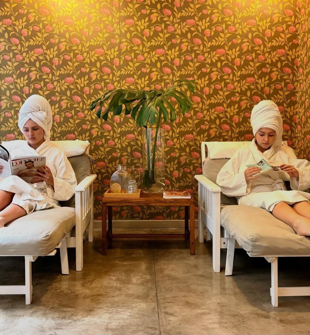 Two individuals seated on white chairs in a room with floral wallpaper, reading magazines, with a potted plant between them.