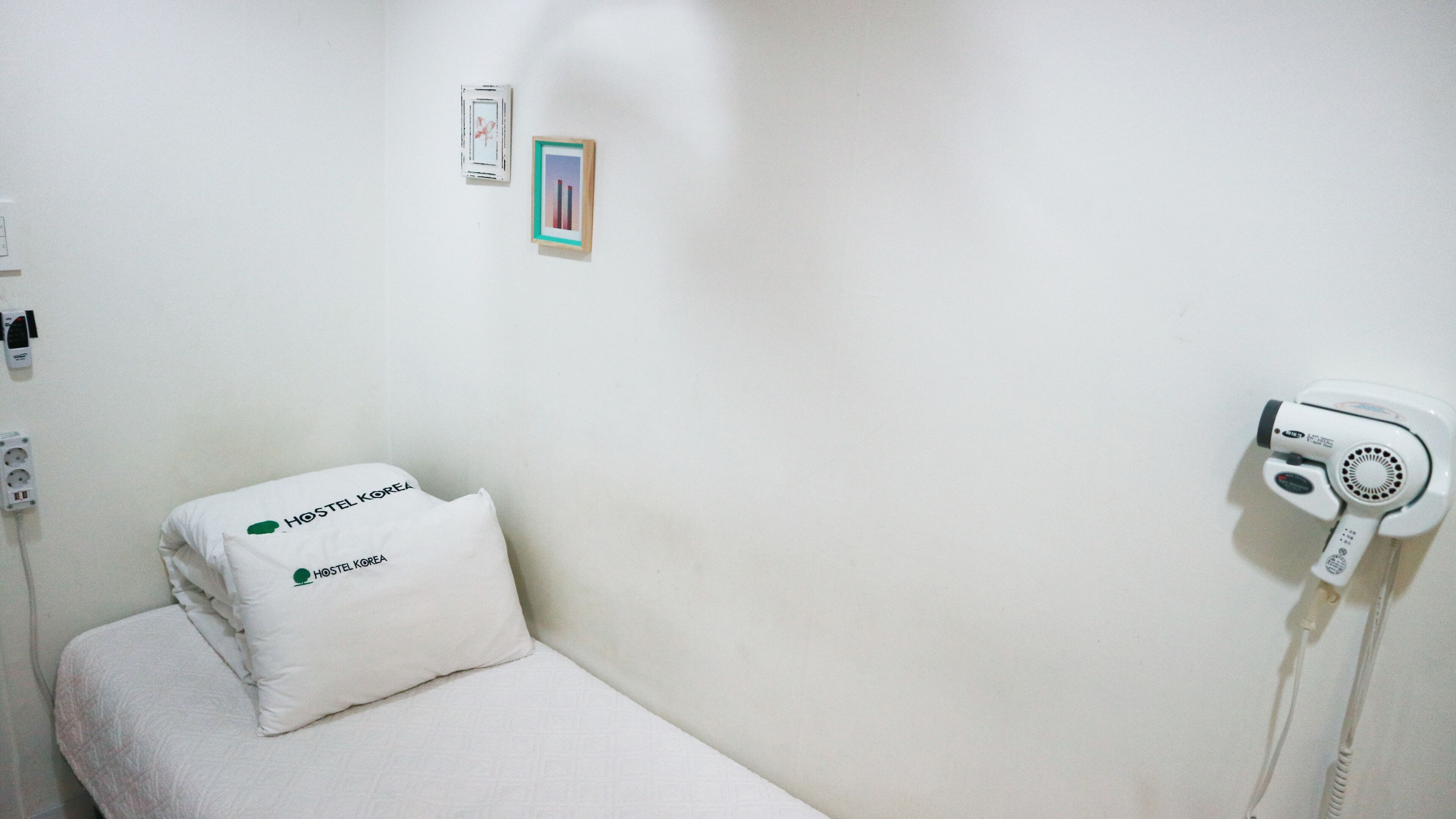 Een schone, minimalistisch ingerichte kamer met een bed, een kussen, een haardroger en een klein fotokader aan de muur.