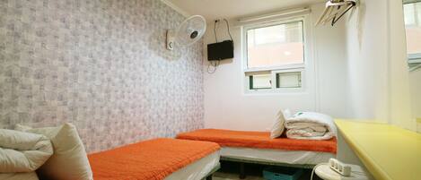 A small, compact room with two single beds, a small table, a chair, a fan, and a window.