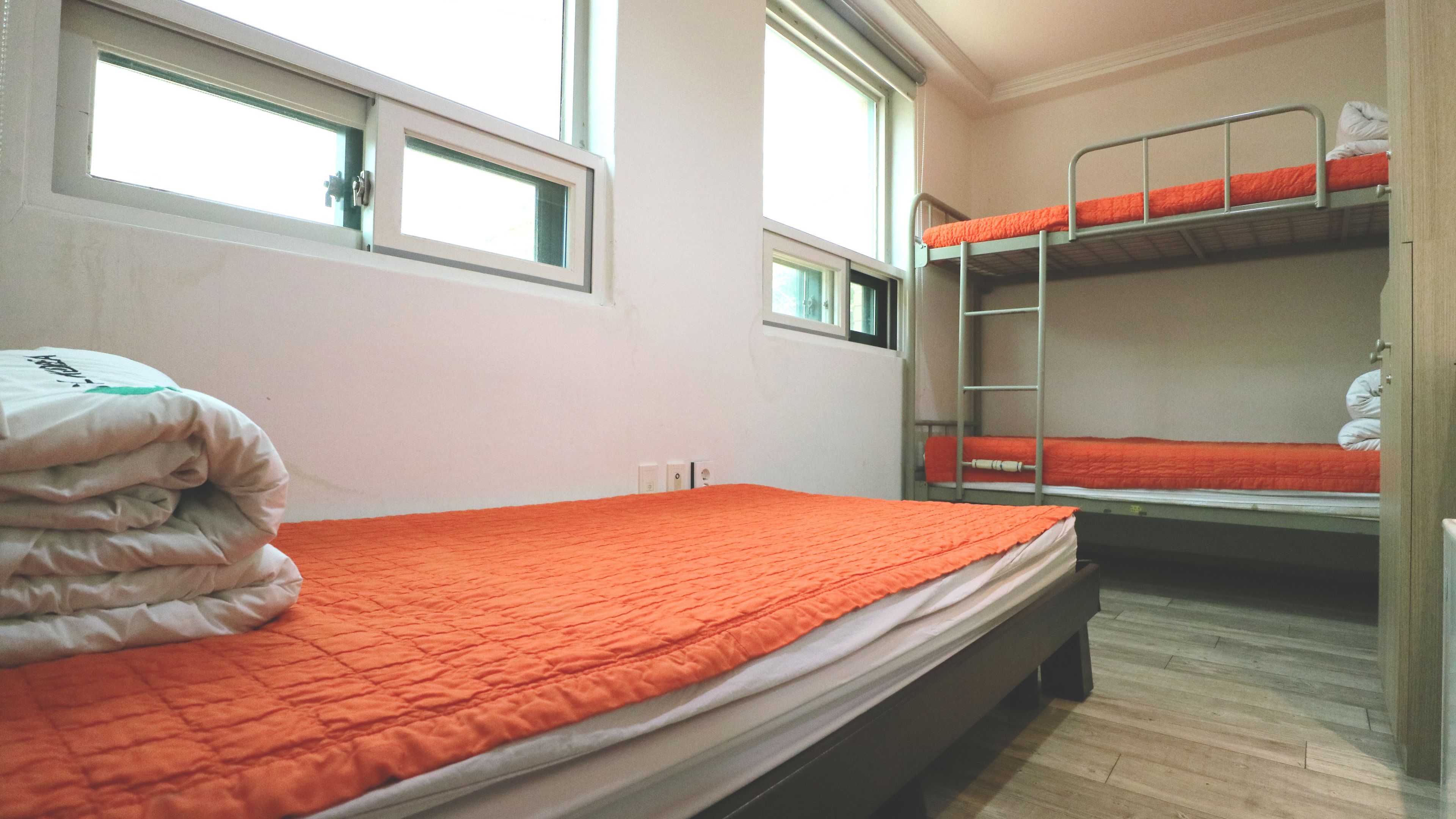 A dormitory room with bunk beds, a bed with an orange blanket, and a window with blinds.