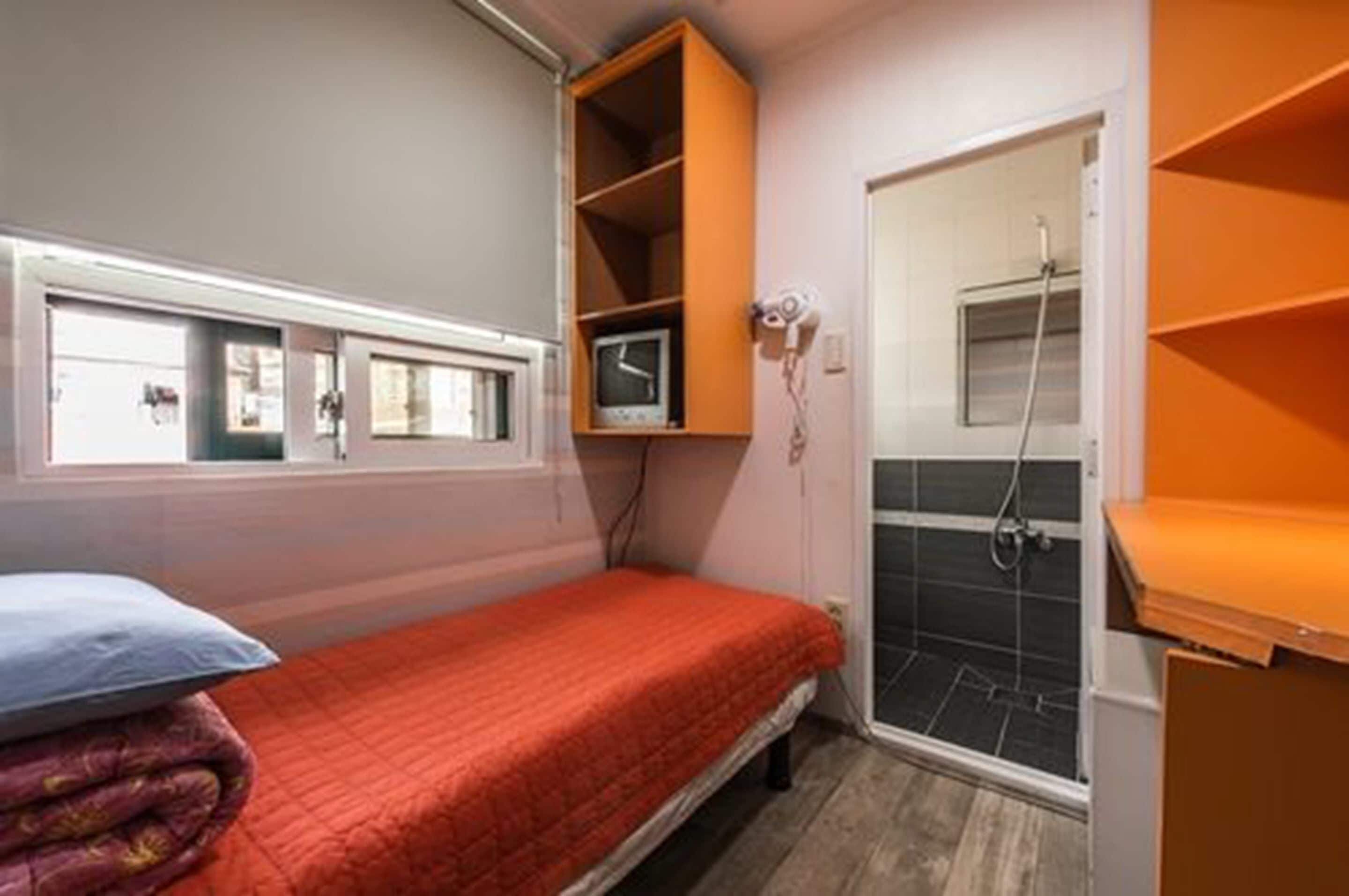 A small, compact room with a single bed, a window, a shower area, and a built-in shelf unit.
