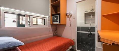 A small, compact room with a single bed, a window, a shower area, and a built-in shelf unit.