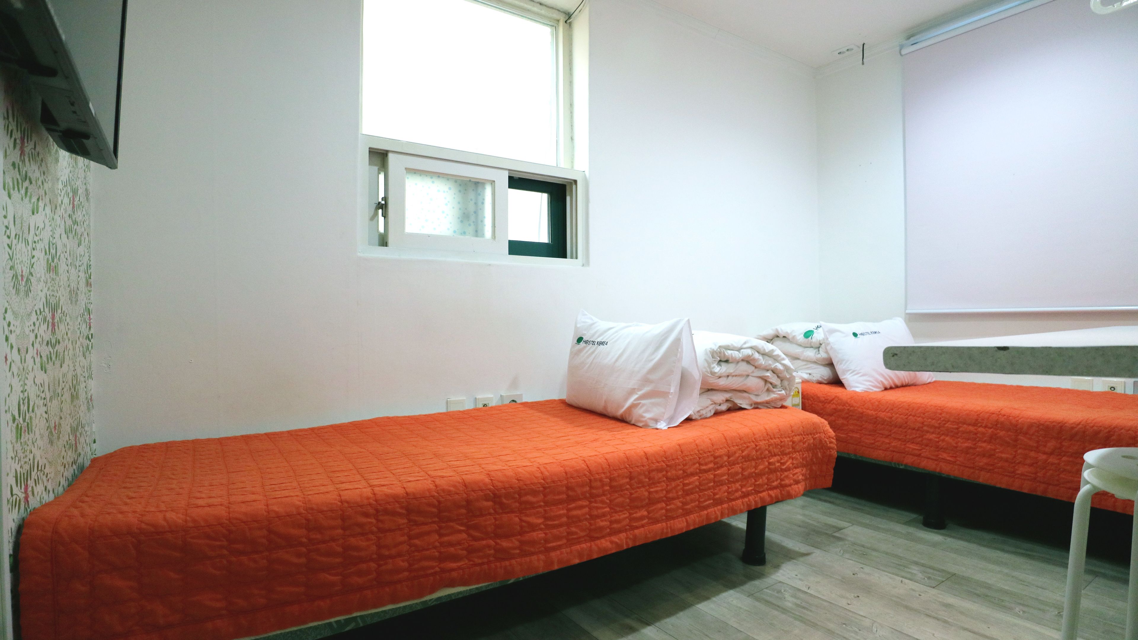 A room with two single beds, each with an orange quilt, white pillows, and a window above one bed.