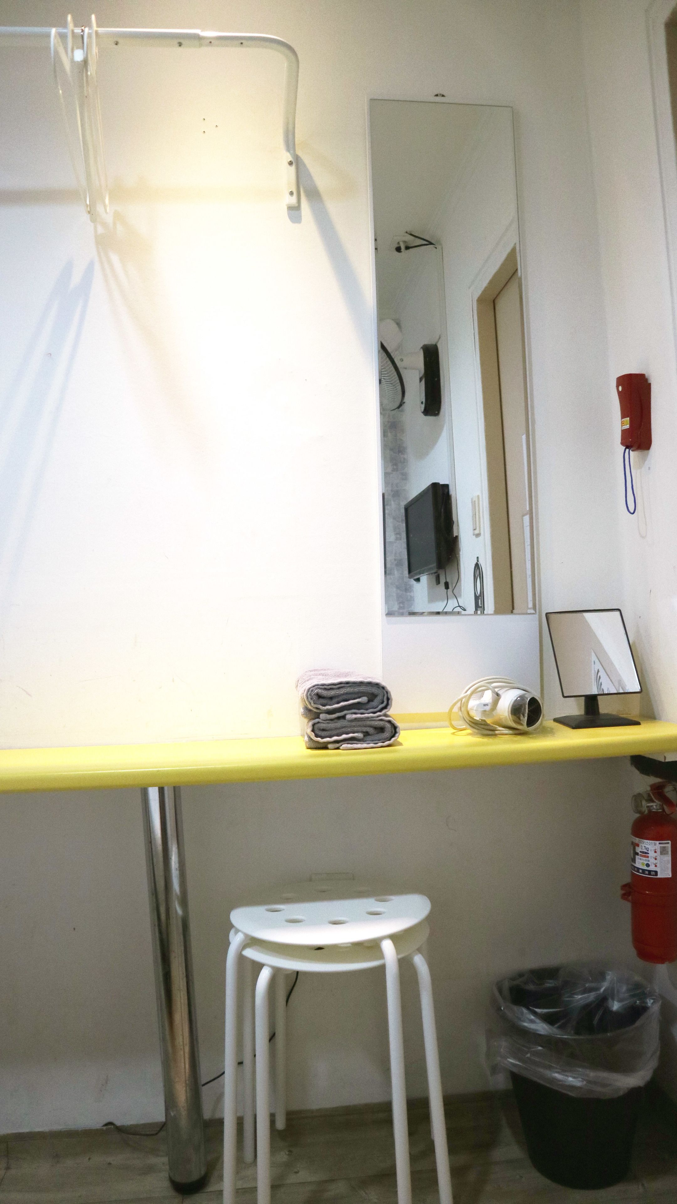 A small room with a yellow desk, a white stool, a mirror, a red telephone, and a black trash bin.