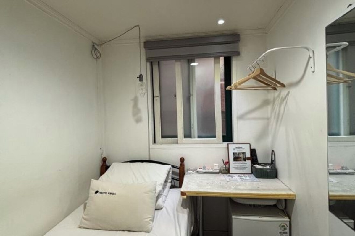 A small, compact room with a single bed, a bedside table, a small desk with a chair, a window with blinds, and a wall-mounted mirror.