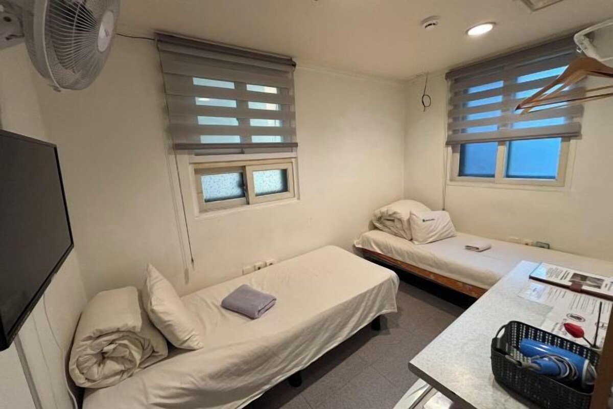 Une petite chambre propre, avec deux lits simples, un bureau avec une chaise, un ventilateur et une fenêtre avec des stores.