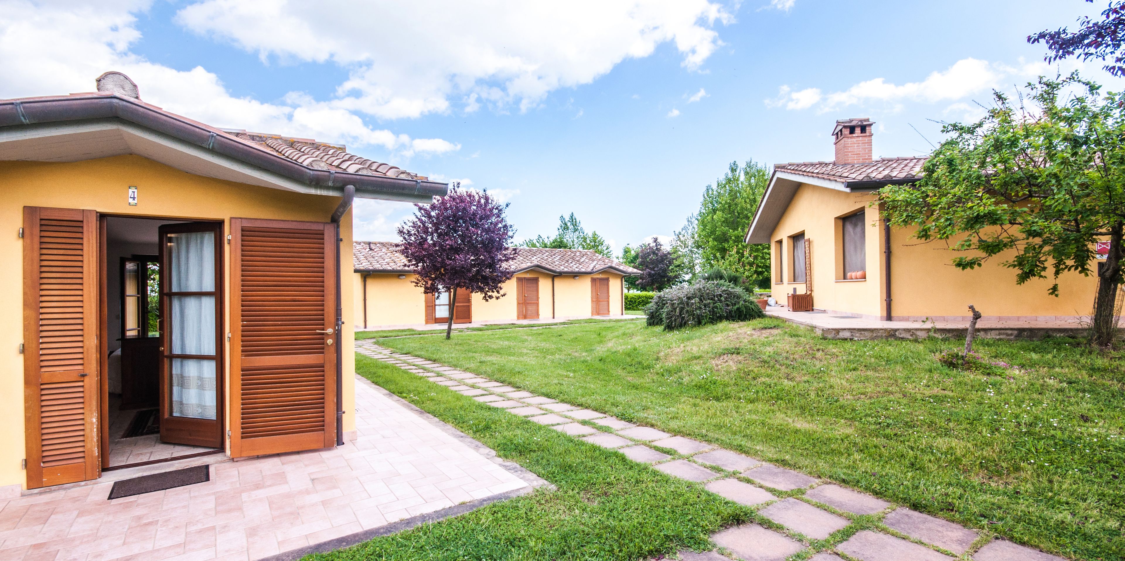A row of yellow buildings with wooden shutters, a paved pathway, and a grassy area with trees.