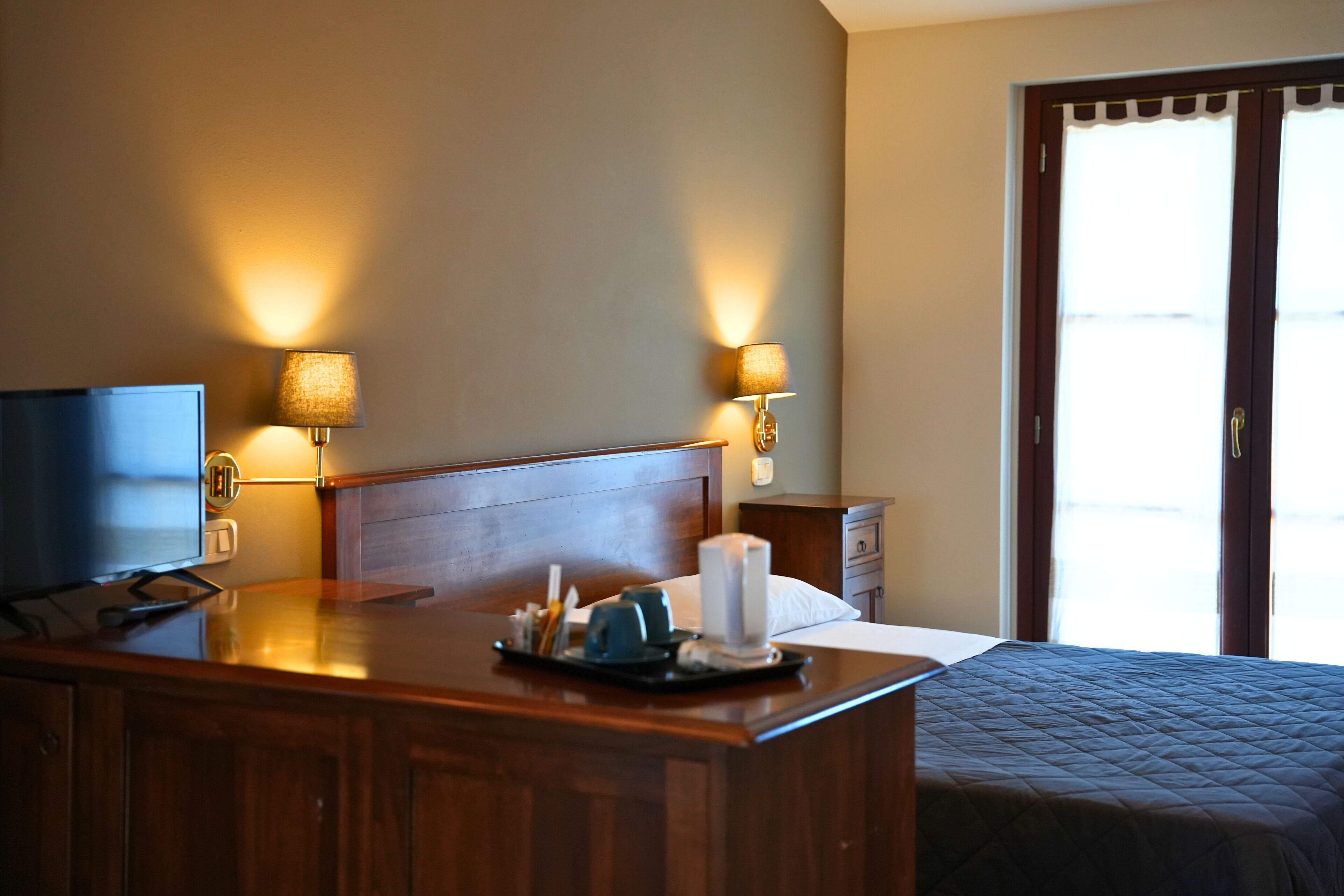 Une chambre d’hôtel avec un bureau en bois, un lit recouvert d’une courtepointe matelassée, deux lampes fixées au mur et un téléviseur placé à gauche.